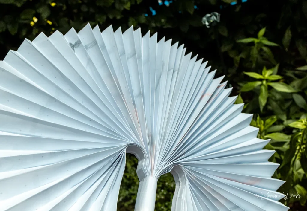 Rueckseite des Flipchart-Sonnenschirms vor gruener Hecke - die weissen Ziehharmonika-Falten bilden einen Halbkreis