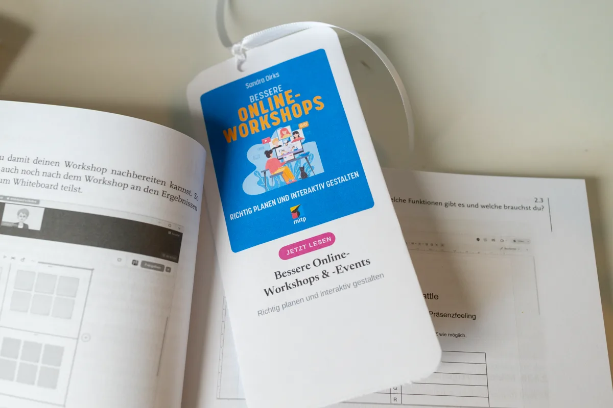 Gefaltetes Lesezeichen mit Mini-Buchcover liegt aufgeklappt auf dem offenen Buch Bessere Online-Workshops