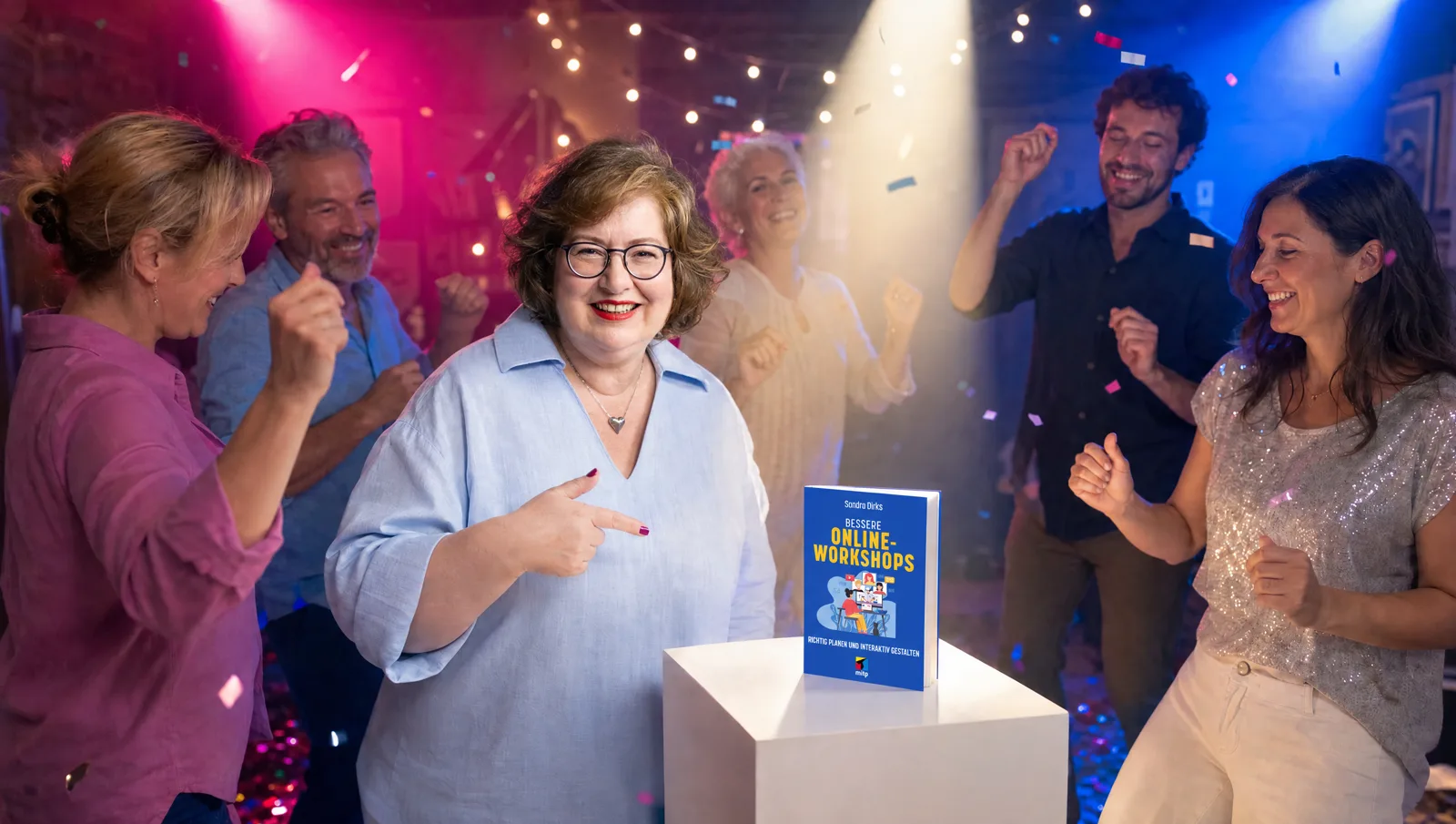 Sandra Dirks tanzt lachend und zeigt auf ihr Buch Bessere Online-Workshops, das im Scheinwerferlicht auf einem weißen Podest steht, um sie herum tanzen Gäste.