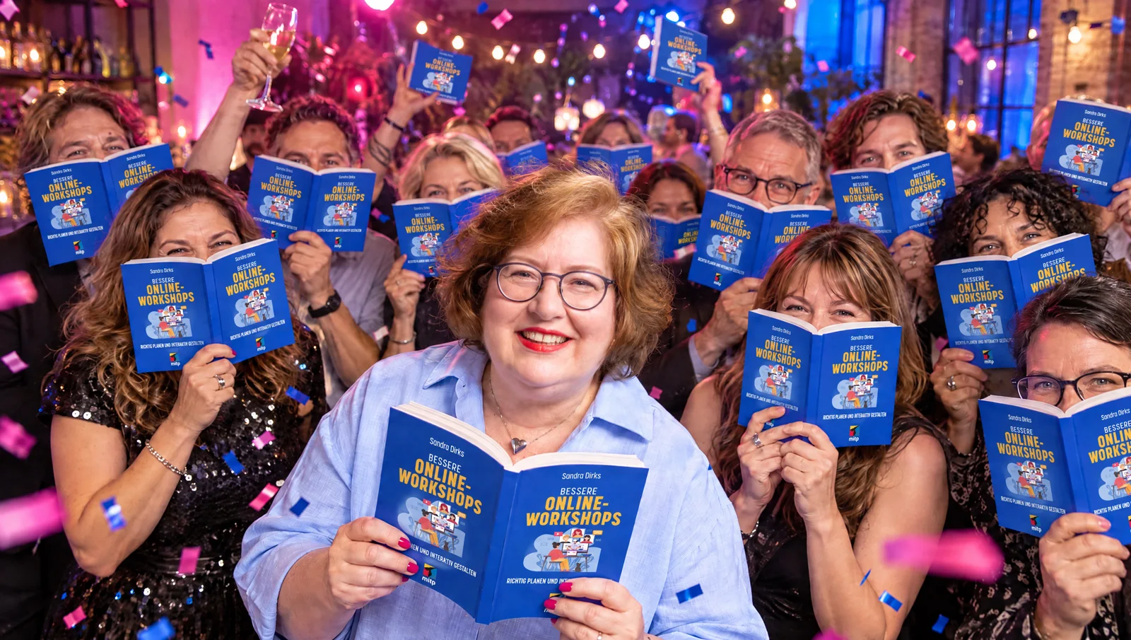 Buchparty: Sandra Dirks steht lachend mit ihrem aufgeschlagenen Buch Bessere Online-Workshops vorn, hinter ihr feiern viele Gäste und halten das Buch hoch, Konfetti fliegt.